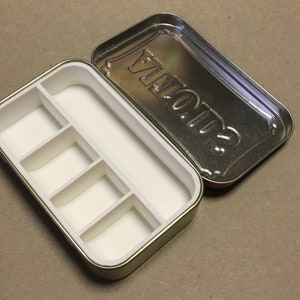 May include: A small, rectangular tin box with a white interior divided into five compartments. The lid of the box is silver and has the word "SUGAR" printed on it.