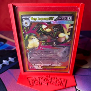 May include: A red 3D-printed Pokémon card display stand holding a Mega Lopunny EX card. The card features a colorful illustration of the Pokémon and game details. The stand has the Pokémon logo at the base.