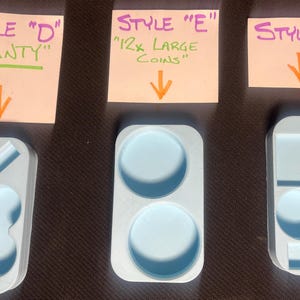 May include: Three silicone molds for resin casting. The molds are labeled "Style D" "SLANTY", "Style E" "12x LARGE COINS", and "Style F". The molds are light blue and have different shapes for casting.