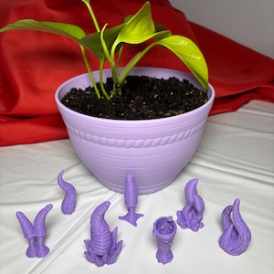 May include: A potted plant with bright green leaves in a lavender pot. Several small, purple, abstract sculptures are arranged in front of the pot. The background is a red fabric.