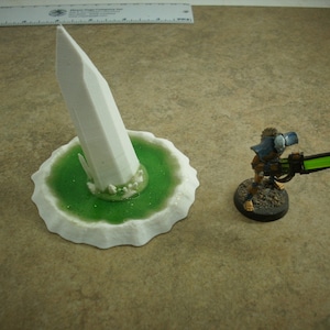 May include: A white 3D printed miniature of a tall, pointed spire on a green base. The spire is surrounded by smaller white crystals. The base is textured and has a wavy edge.