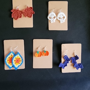 May include: Five pairs of crocheted Halloween earrings displayed on brown card stock. The earrings are in the shapes of leaves, skulls, pumpkins, bats, and a geometric design.