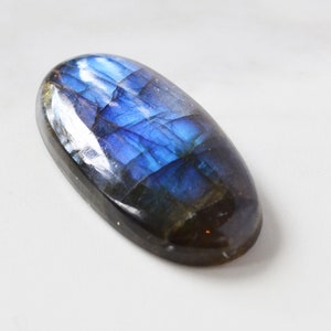 Pode incluir: Uma pedra preciosa de labradorita em forma oval com uma superf&iacute;cie polida. A pedra exibe um vibrante jogo de cores, com azuis e roxos iridescentes. A gema &eacute; colocada sobre um fundo branco, real&ccedil;ando sua beleza natural.