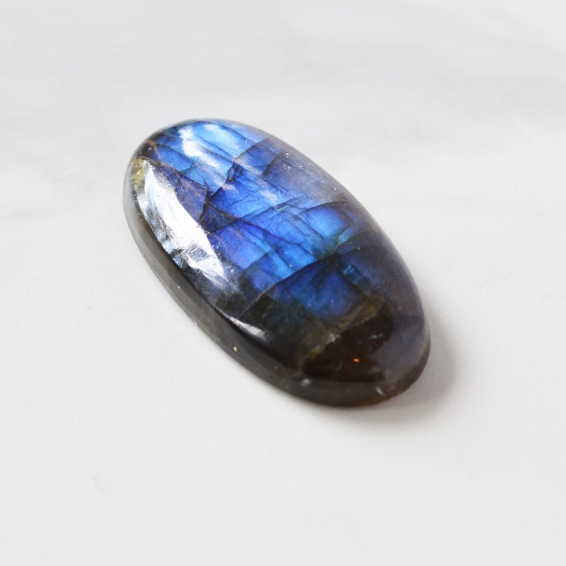 Pode incluir: Uma pedra preciosa de labradorita em forma oval com uma superf&iacute;cie polida. A pedra exibe um vibrante jogo de cores, com azuis e roxos iridescentes. A gema &eacute; colocada sobre um fundo branco, real&ccedil;ando sua beleza natural.