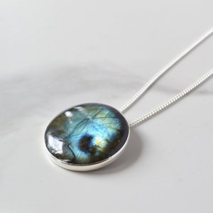 Pode incluir: Um pendente redondo de labradorite com moldura de prata, suspenso por uma delicada corrente de prata. A pedra preciosa exibe um jogo de cores cativante, com tons de azul, verde e dourado. O colar &eacute; apresentado numa superf&iacute;cie branca.