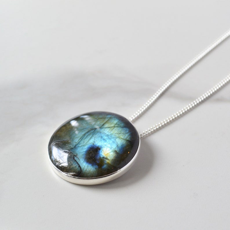 Pode incluir: Um pendente redondo de labradorite com moldura de prata, suspenso por uma delicada corrente de prata. A pedra preciosa exibe um jogo de cores cativante, com tons de azul, verde e dourado. O colar &eacute; apresentado numa superf&iacute;cie branca.