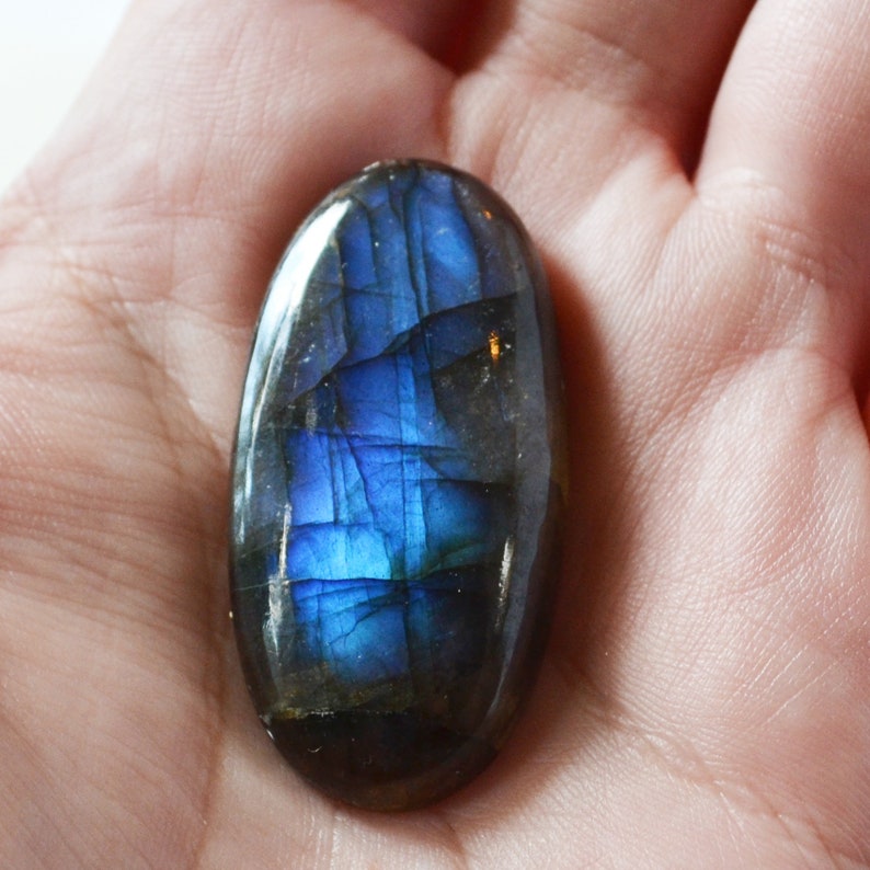 Pode incluir: Uma pedra preciosa de labradorita em forma oval com uma superf&iacute;cie polida. A pedra exibe um brilho azul vibrante e iridescente, com tons cinza e preto mais escuros. A pedra preciosa &eacute; segurada em uma m&atilde;o.