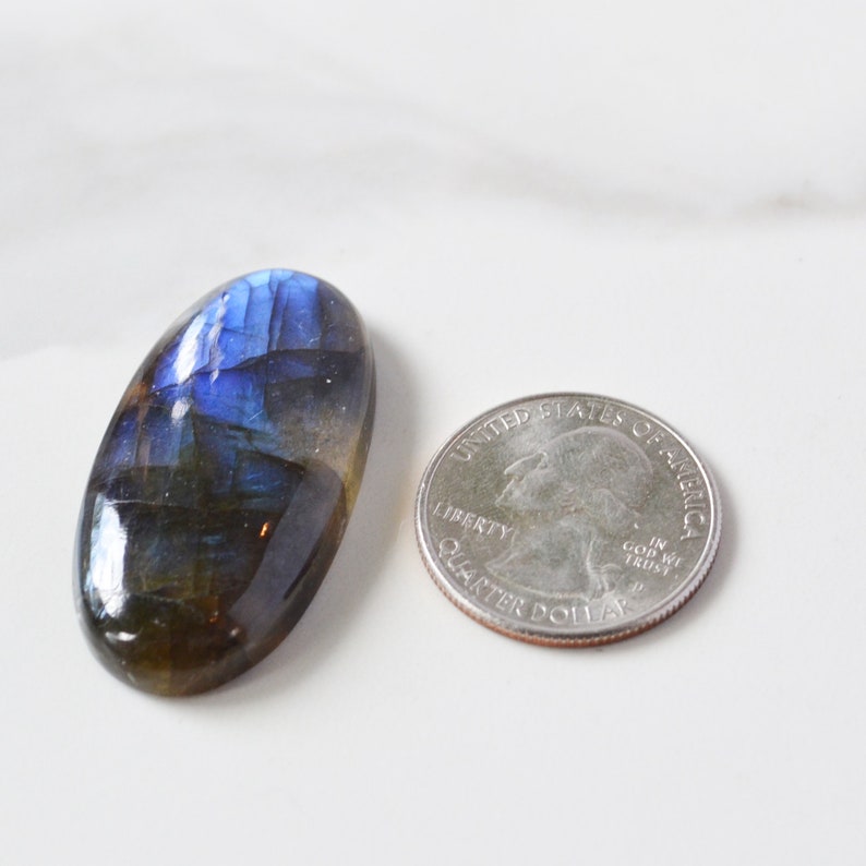 Pode incluir: Pedra preciosa de labradorita oval com flashes iridescentes azuis e verdes. A pedra polida &eacute; exibida ao lado de uma moeda para compara&ccedil;&atilde;o de tamanho. A pedra preciosa tem aproximadamente 3,2 cm de comprimento.