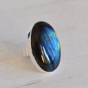 Pode incluir: Um anel de prata com uma grande pedra de labradorita oval. A pedra exibe um jogo de cores brilhante, com tons de azul, verde e amarelo. O anel tem uma banda simples e polida.