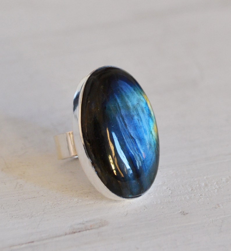 Pode incluir: Um anel de prata com uma grande pedra de labradorita oval. A pedra exibe um jogo de cores brilhante, com tons de azul, verde e amarelo. O anel tem uma banda simples e polida.