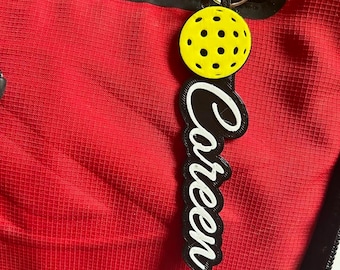 Pode incluir: Um chaveiro preto e branco com a palavra "Careen" escrita em itálico. O chaveiro apresenta um mini pickleball amarelo e preto.