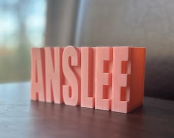 May include: A coral-coloured, 3D-printed block with the name "ANSLEE" in large, block letters. The letters are evenly spaced and stand upright, creating a personalised decorative item. The object is sitting on a dark wooden surface.