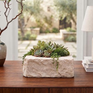 May include: A rectangular planter carved from light beige natural stone, filled with a variety of green succulents including echeveria, sedum, and string of pearls. The planter has a rough, textured exterior surface and sits on a wooden surface.