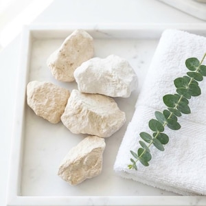 May include: A square white marble tray containing four irregular beige stones, a folded white cotton towel, and a single green eucalyptus sprig with small round leaves. The tray has a smooth, polished surface with subtle grey veining.