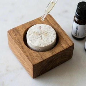 May include: A square wooden block with a circular cutout containing a porous, off-white stone disc. A glass dropper releases liquid onto the disc. Three small amber glass bottles with black caps are labeled Lavender, Bergamot, and Patchouli.