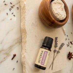 May include: A 10ml amber glass bottle of dōTERRA Lavender essential oil with a black cap, positioned next to two sprigs of dried lavender, a small wooden bowl containing a beige powder, and scattered dried lavender buds and a star anise.