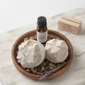 May include: A round wooden tray containing two rough, off-white limestone stones, a small amber glass bottle labeled Lavender & Bergamot essential oil, a sprig of dried lavender, and small pebbles. A wooden block is engraved Natural Limestone Diffuser.