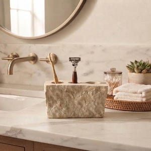 May include: A rectangular bathroom organizer carved from light beige natural stone with a rough, textured exterior. The piece features three circular openings on the top surface, currently holding a wooden toothbrush and a metal razor.