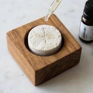 May include: A square wooden block with a circular center cutout holding a round, porous, off-white stone diffuser. A glass dropper dispenses liquid onto the stone. Three small dark glass bottles labeled Lavender, Bergamot, and Patchouli sit nearby.