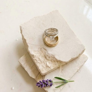 May include: A stack of three rings consisting of a silver band with a square-cut center stone, a silver band with small pavé-set stones, and a plain gold band. A single sprig of purple lavender rests on the textured stone surface below the rings.