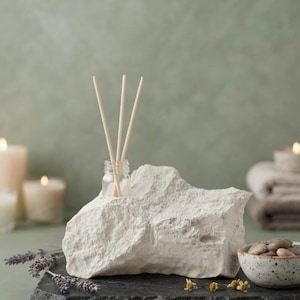 May include: A white, rough-textured stone diffuser base with a small glass bottle inserted into the top. Three light-colored wooden reeds extend upward from the bottle. The stone sits on a dark, flat slate surface alongside small pebbles.