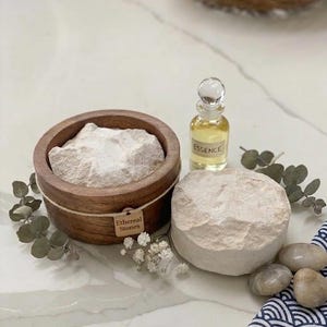 May include: A round wooden bowl filled with white powder, featuring a tag labeled Ethereal Stones. Beside it sits a small glass bottle of yellow liquid labeled ESSENCE, a round textured white stone, small white flowers, and smooth gray pebbles.