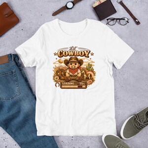 Könnte beinhalten: Weißes T-Shirt mit einem Cartoon-Baby-Cowboy-Design. Das Baby trägt einen braunen Hut und ein rotes Halstuch, mit dem Text "Lil' Cowboy" darüber und "LOADING... 75%" darunter. Das Design umfasst einen Kaktus, ein Hufeisen und Blumen, alles in Sepia-Tönen.
