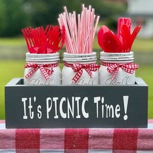 May include: A gray wooden box with the text "It's Picnic Time!" painted on the front. The box holds three white mason jars with red and white gingham ribbon. The jars are filled with red plastic forks, red plastic spoons, and red and white striped straws.