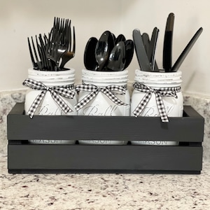 May include: Three white mason jars with black and white checkered ribbon bows, holding black plastic utensils. The jars are sitting in a black wooden crate.