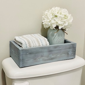 May include: A gray wooden box with a white towel and a white flower arrangement in a mason jar labeled "Ball" on top of a white toilet tank.