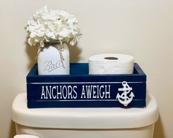 May include: A blue wooden box with the text "Anchors Aweigh" and a white anchor. The box is on top of a white toilet and holds a roll of toilet paper and a white mason jar with white flowers.