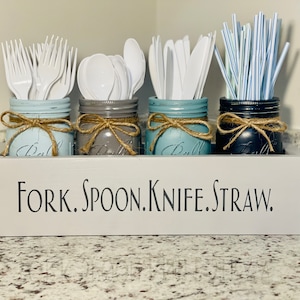 May include: Four mason jars with twine and bows, painted in different colors, are used to hold forks, spoons, knives, and straws. The jars are sitting in a wooden box with the words "FORK. SPOON. KNIFE. STRAW." painted on the front.