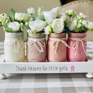 May include: Four mason jars painted in shades of pink and white, filled with white flowers, are arranged on a white wooden tray with the text "Thank Heaven for little girls" and a pink flower graphic.