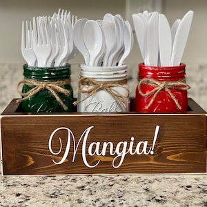 May include: A wooden box with the word "Mangia!" on the front. The box holds three mason jars, each painted a different color: green, white, and red. The jars are filled with plastic utensils, including forks, spoons, and knives.