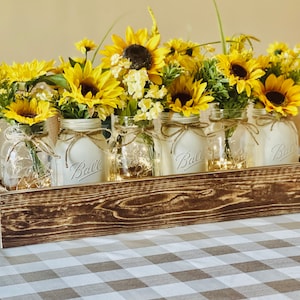 May include: A rustic wooden box filled with mason jars, each containing a bouquet of yellow sunflowers. The jars are tied with twine and have the word "Ball" printed on them.
