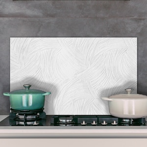 Op de afbeelding: Een witte keukenachterwand met een textuurpatroon. Twee pannen, een groene en een witte, staan op een zwart fornuis.