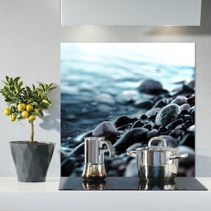 Puede incluir: Una placa de cocción de acero inoxidable con una superficie de vidrio negro. Una olla de plata y una cafetera de plata están en la placa de cocción. El fondo es una imagen borrosa de una playa con rocas y agua.