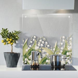 May include: A kitchen setting with a clear glass splashback displaying a floral design of white lily of the valley flowers and green leaves. A stainless steel pot and espresso maker sit on a black hob. A potted lemon tree is on the left.