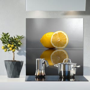 May include: A stainless steel hob with a black glass cooktop. A silver saucepan with a glass lid and a silver coffee maker are on the hob. A lemon cut in half and a whole lemon are on a silver splashback behind the hob. A small potted citrus tree with green leaves and yellow fruit is on the counter to the left of the hob.