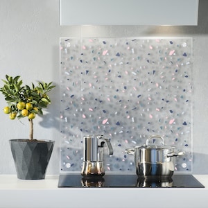 Könnte beinhalten: Eine Küche mit einer weißen Arbeitsplatte und einem Edelstahlkochfeld. Ein kleiner Topf-Zitrusbaum steht auf der Theke. Die Rückwand ist eine durchsichtige Glasscheibe mit einem Terrazzo-Muster in den Farben Rosa, Blau und Grau. Eine silberne Kaffeekanne und ein silberner Topf stehen auf dem Kochfeld.