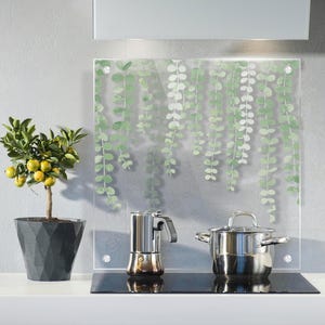 Könnte beinhalten: Küchenszene mit einer Glas-Rückwand mit grünem Blattmuster. Links steht ein kleiner Topf mit Zitrusfrüchten mit gelben Früchten. Ein Edelstahl-Topf, eine Espressomaschine und ein schwarzes Kochfeld sind ebenfalls zu sehen.