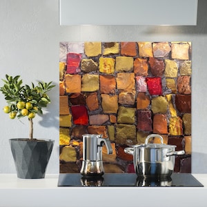 May include: A kitchen backsplash with a mosaic tile design in shades of brown, red, and gold. A silver coffee pot and a silver saucepan are on a black stovetop.
