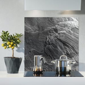 May include: A kitchen scene featuring a small potted lemon tree with yellow fruit, a stainless steel stovetop, a silver espresso maker, and a stainless steel pot. A large black and gray stone-like backsplash is behind the stovetop.