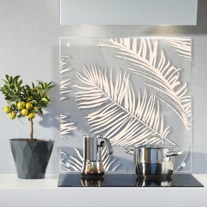 Könnte beinhalten: Eine transparente Küchenrückwand aus Glas mit einem cremefarbenen Palmenblattmuster. Ein Kochtopf aus Edelstahl und eine Espressomaschine stehen auf einem schwarzen Kochfeld. Ein kleiner Topf mit Zitrusfrüchten mit gelben Früchten steht auf der Theke.