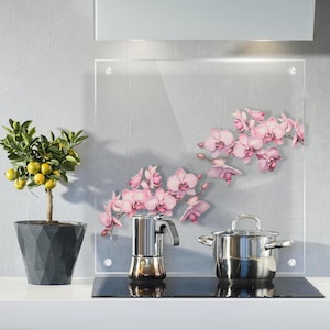 May include: A kitchen setting with a small potted citrus tree bearing yellow fruit, a stainless steel espresso maker, and a stainless steel saucepan. A clear glass panel with pink orchid decals is mounted on the wall above the hob.