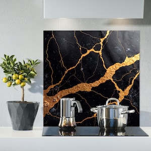 May include: A black and gold marble-patterned kitchen backsplash with a stainless steel pot and espresso maker on a black stovetop. A small potted lemon tree with yellow fruit sits to the left. The backsplash has gold veining.