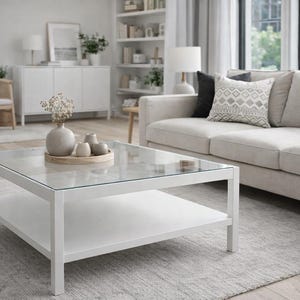 Puede incluir: Salón moderno con una mesa de centro blanca con tapa de cristal y estante inferior. Un sofá beige con cojines decorativos descansa sobre una alfombra gris. Jarrones decorativos y una bandeja están sobre la mesa de centro. Una silla de madera y un armario blanco están al fondo.
