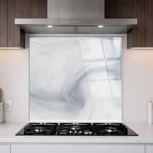 May include: A square kitchen splashback panel featuring a swirling, abstract marble-like pattern in shades of white and soft grey. The panel is positioned vertically behind a black gas hob, set against a light-coloured kitchen wall.