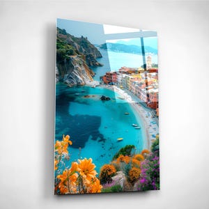 Puede incluir: Una vibrante impresión de un pueblo costero con edificios coloridos, una playa de arena y agua turquesa. La imagen presenta una vista del mar, barcos y exuberante vegetación, con flores de color naranja brillante en primer plano. La obra se exhibe sobre una superficie brillante.