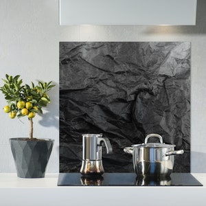 May include: A black and white crumpled paper design kitchen backsplash with a silver pot and a silver coffee maker on a black electric hob.